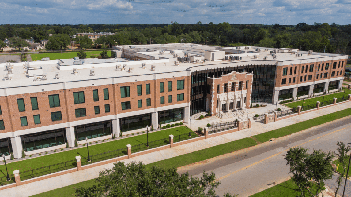 Aerial view of a large, modern three-story Learning Community building with a mix of brick and glass exterior, set along a tree-lined street with sidewalks and green lawns.