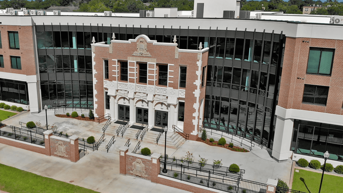 A modern three-story school building with large glass windows and a historic central entrance, this Living and Learning Community features detailed stonework, three arched doorways, lush landscaping, and a welcoming paved walkway.