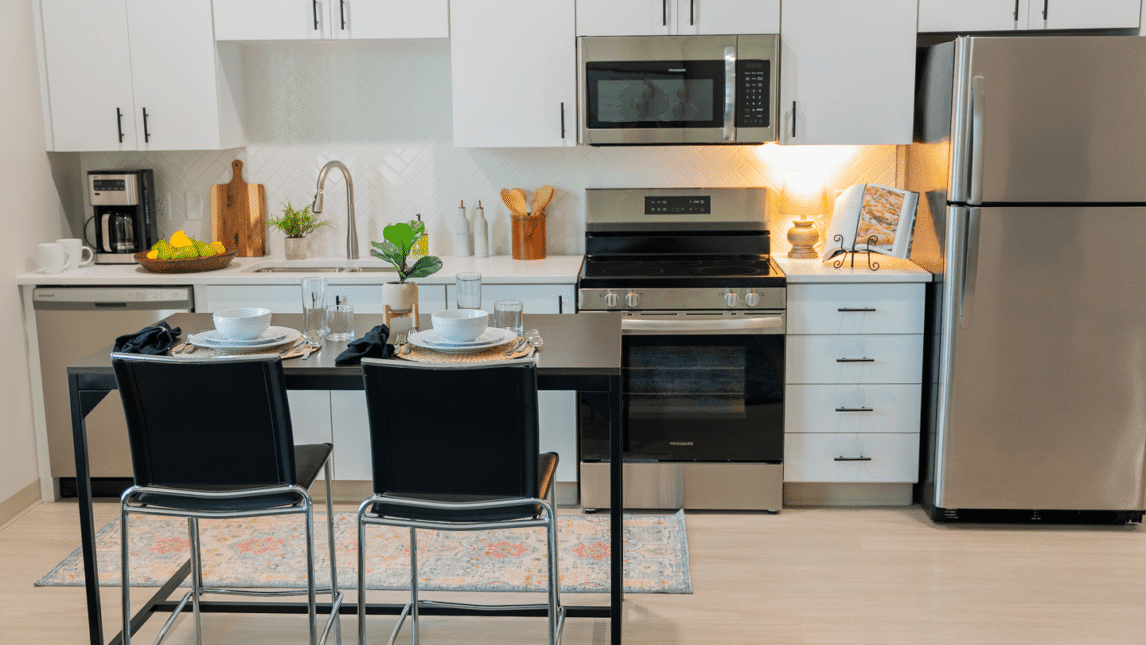 Modern kitchen with white cabinets, stainless steel appliances, and a small dining table for two with black chairs—perfect for a Living & Learning Community setting. Countertop features kitchen essentials, fresh fruit, and stylish decor items.