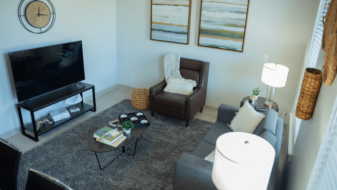 A modern Living & Learning Community living room with a gray sofa, brown armchair, coffee table with books and decor, TV on a stand, abstract paintings, wall clock, and floor lamp on a gray rug with natural light from a window.