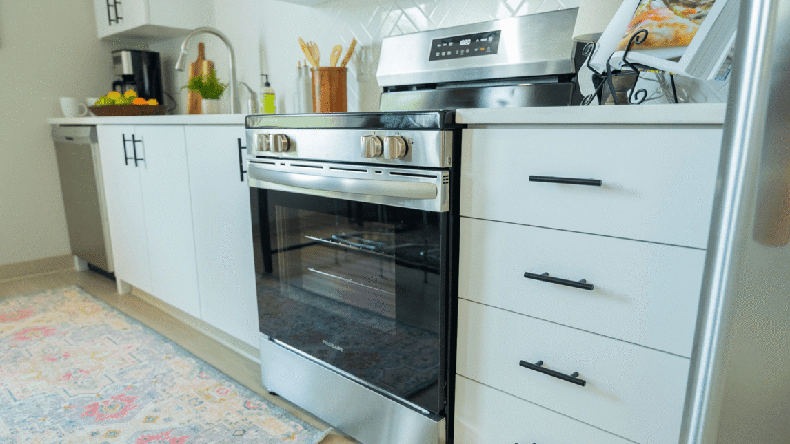 Modern kitchen with a stainless steel oven and white cabinets in a vibrant Living & Learning Community. Kitchen utensils, plants, and a bowl of lemons sit on the counter, while a colorful rug brightens the floor in front of the oven.