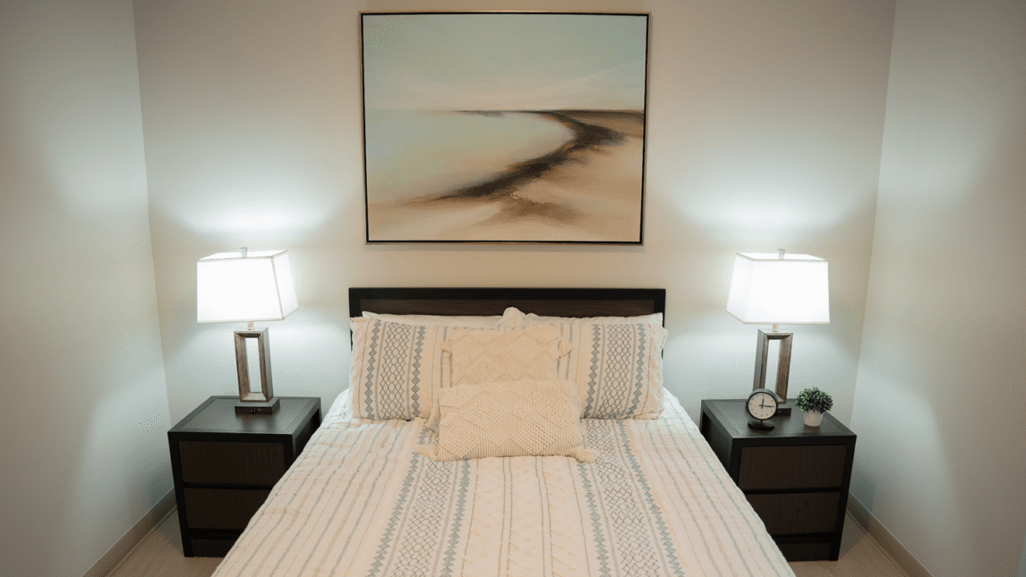 A neatly made bed with patterned bedding is centered between two dark nightstands, each with a lamp. Above the bed hangs an abstract landscape painting, creating a cozy Living & Learning Community vibe. An alarm clock and small plant sit on the right nightstand.