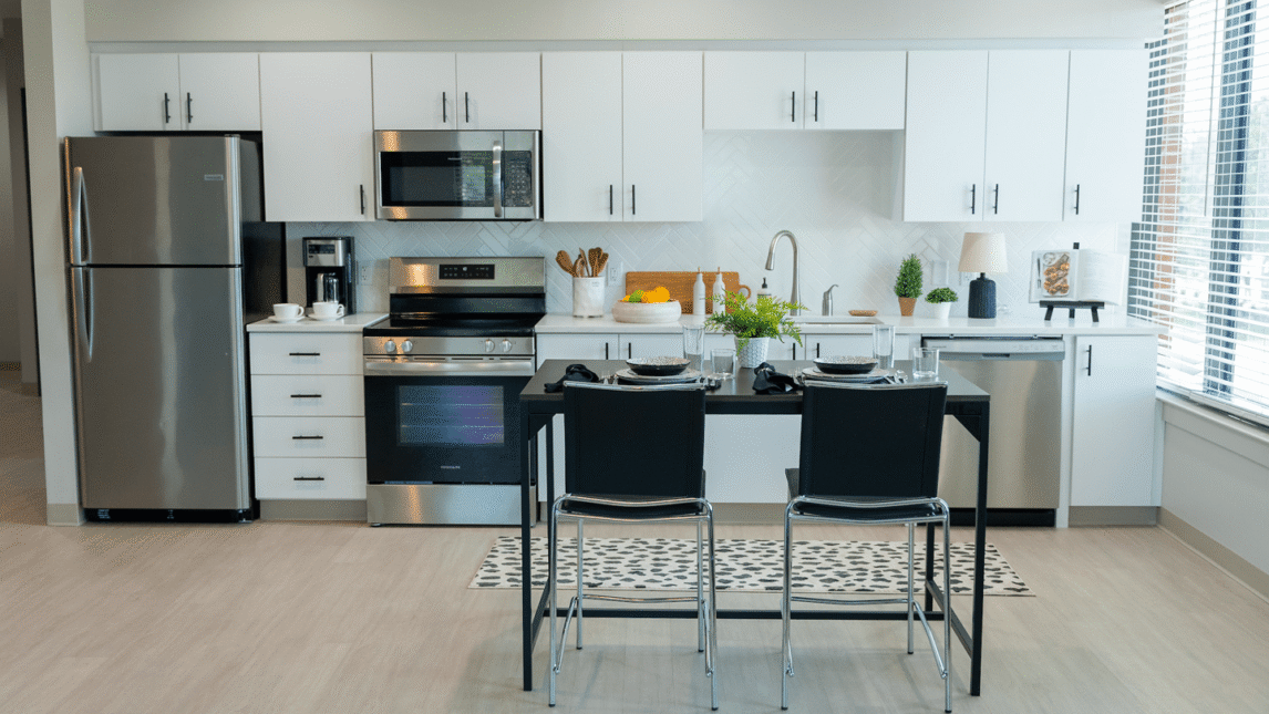 Modern kitchen with white cabinets, stainless steel appliances, a sink, countertop decor, and a small dining table with two black chairs—ideal for a Living & Learning Community. Large windows on the right let in natural light.