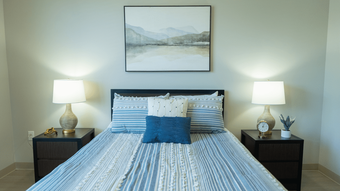 A neatly made bed with blue and white striped bedding, two pillows, and a dark blue accent pillow creates a welcoming Living and Learning Community vibe. Matching lamps on dark bedside tables frame a landscape painting above the bed.