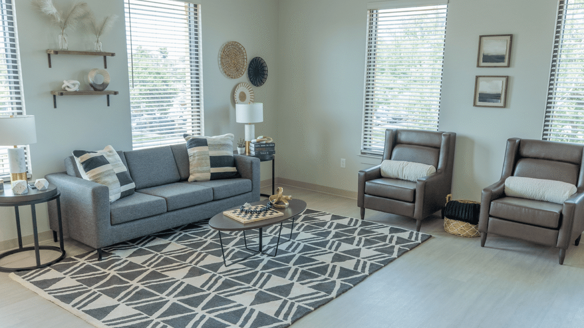Bright living room with a gray sofa, two brown armchairs, a geometric rug, a coffee table with a chess set—perfect for gathering in this Living & Learning Community—plus wall art, shelves, and large windows with blinds letting in natural light.