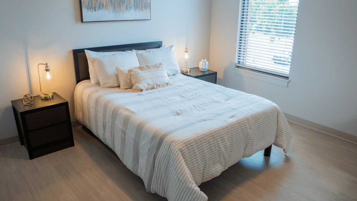 A neatly made bed with white striped bedding and decorative pillows sits between two dark nightstands with lamps, creating an inviting space in this bright Living & Learning Community bedroom with light wood floors and a window with blinds.