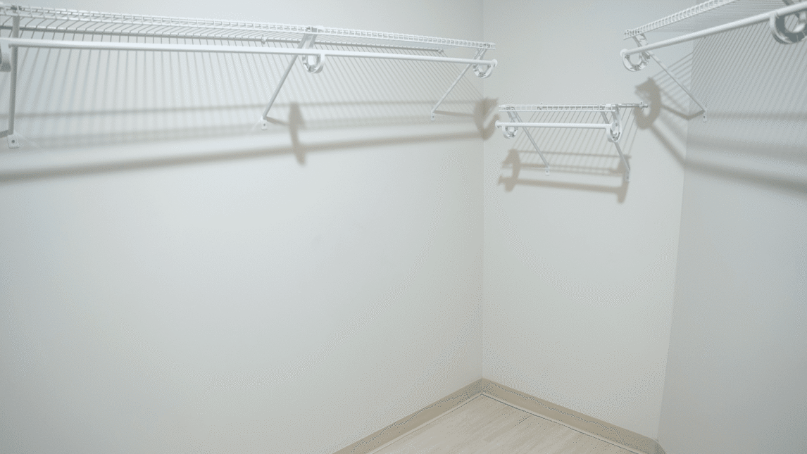 Empty walk-in closet with beige walls and light wood flooring, featuring white wire shelves and hanging rods along three walls—an ideal storage space for any Living & Learning Community. The area is well-lit, clean, and uncluttered.