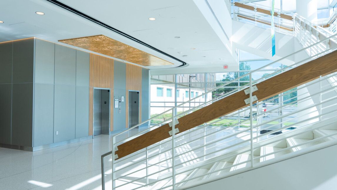 Bright, modern building interior with white railings, wooden accents, and large windows letting in natural light; the Trauma Tower elevator area is visible on the left, and a staircase ascends on the right. Bright, modern building interior with white railings, wooden accents, and large windows letting in natural light; the Trauma Tower elevator area is visible on the left, and a staircase ascends on the right.