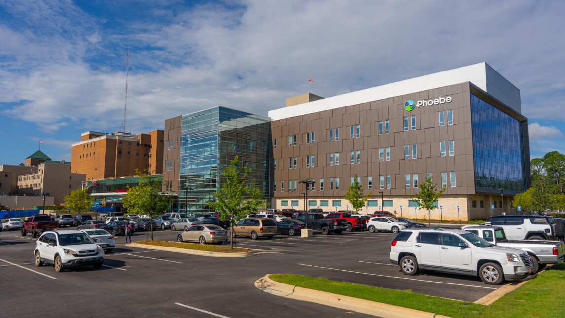 Modern hospital tower with large glass windows and Phoebe logo, surrounded by a parking lot filled with cars, under a blue sky. The facility specializes in trauma and critical care services. Modern hospital tower with large glass windows and Phoebe logo, surrounded by a parking lot filled with cars, under a blue sky. The facility specializes in trauma and critical care services.