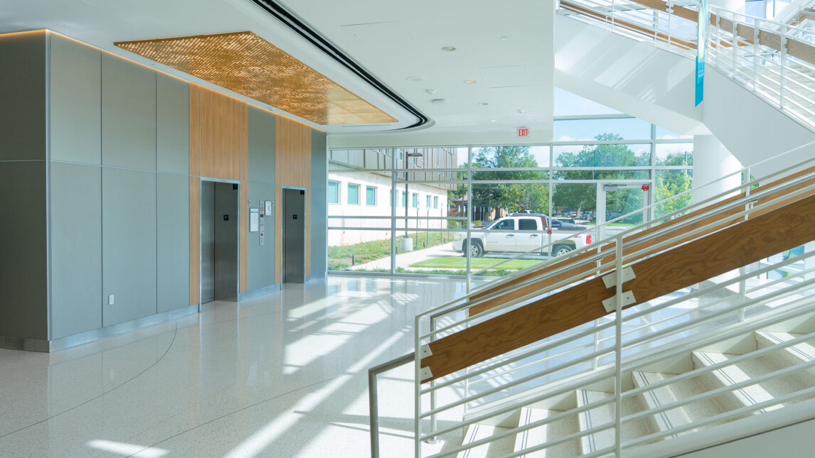 Modern building lobby in the Critical Care Tower with large glass windows, two elevators, a staircase with wooden handrails, and sunlight streaming in. A white truck and trees are visible outside through the windows. Modern building lobby in the Critical Care Tower with large glass windows, two elevators, a staircase with wooden handrails, and sunlight streaming in. A white truck and trees are visible outside through the windows.