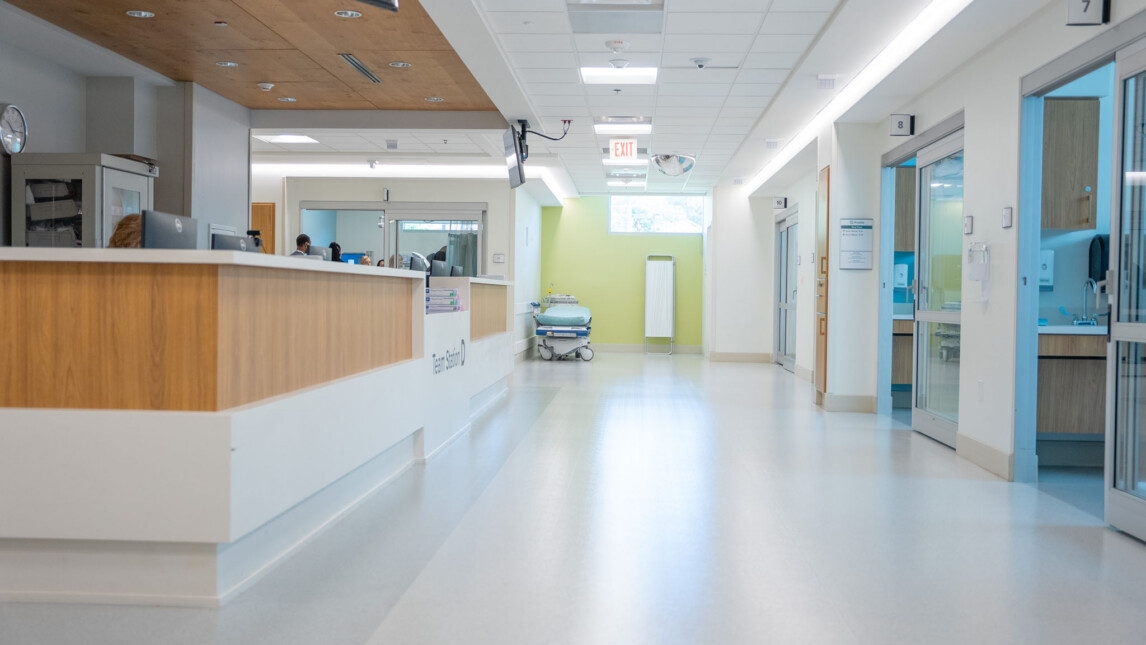 A bright, clean hospital hallway with a wooden reception desk on the left, patient rooms on the right, and an empty gurney near a window at the end of the hall in the Trauma Tower. The space appears modern and quiet. A bright, clean hospital hallway with a wooden reception desk on the left, patient rooms on the right, and an empty gurney near a window at the end of the hall in the Trauma Tower. The space appears modern and quiet.
