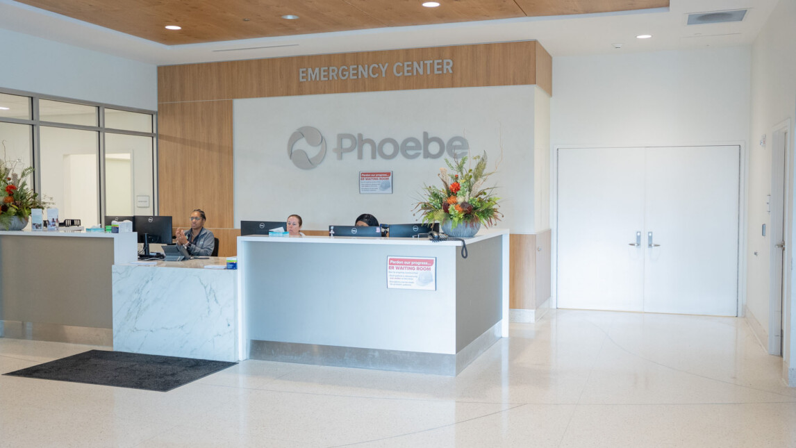 Modern hospital emergency center reception area with three staff members behind the counter, large floral arrangements, and prominent Phoebe logo on the back wall. This clean, brightly lit space supports trauma and critical care needs. Modern hospital emergency center reception area with three staff members behind the counter, large floral arrangements, and prominent Phoebe logo on the back wall. This clean, brightly lit space supports trauma and critical care needs.