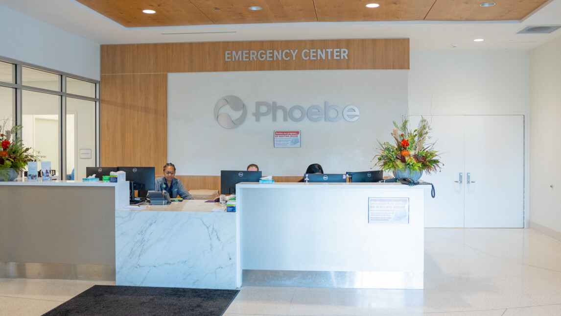 Reception area of an emergency center with two staff members seated behind a marble counter, flower arrangements on both ends, and a sign reading Phoebe on the wall behind them—setting the stage for trauma and critical care services. Reception area of an emergency center with two staff members seated behind a marble counter, flower arrangements on both ends, and a sign reading Phoebe on the wall behind them—setting the stage for trauma and critical care services.