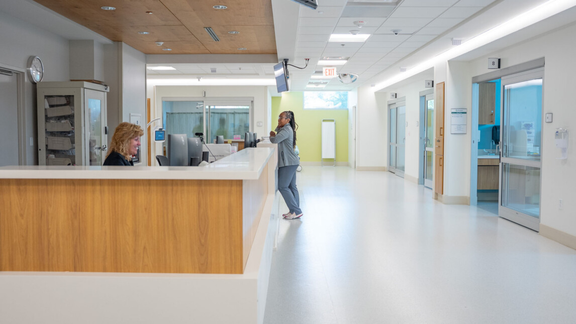 A woman speaks to a receptionist at a hospital front desk in a bright, clean hallway, with medical rooms and critical care equipment visible in the background. A woman speaks to a receptionist at a hospital front desk in a bright, clean hallway, with medical rooms and critical care equipment visible in the background.