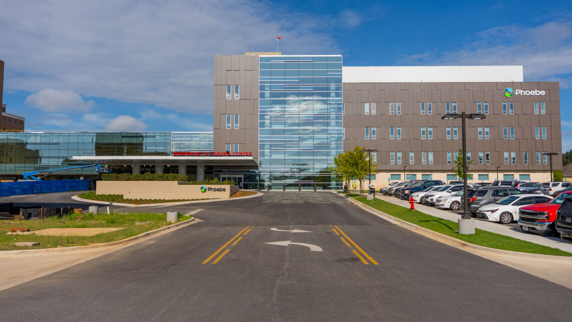 A modern hospital tower with a glass facade and Phoebe signs, surrounded by parked cars and greenery, under a partly cloudy sky. The main entrance, leading to critical care and trauma services, is visible at the end of a wide, paved road. A modern hospital tower with a glass facade and Phoebe signs, surrounded by parked cars and greenery, under a partly cloudy sky. The main entrance, leading to critical care and trauma services, is visible at the end of a wide, paved road.