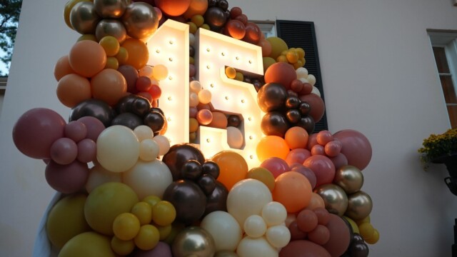 A large, illuminated number 15 is surrounded by a colorful arrangement of balloons in yellow, orange, brown, gold, and pink, celebrating the Annual Banquet of Phoebe Physicians Group outside a light-colored house.