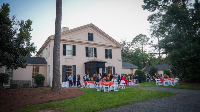People gather outside a large, light-colored house with black shutters for the Phoebe Physicians Group Annual Banquet. White tables and chairs with red tablecloths are set up on the lawn, surrounded by tall trees and greenery.