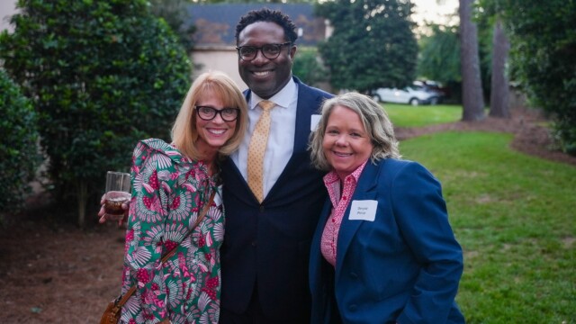 Three people smile outdoors at the Annual Banquet. The woman on the left holds a drink, the man in the center wears a suit and tie, and the woman on the right—wearing a name tag—represents Phoebe Physicians Group. Trees and grass create a scenic backdrop.