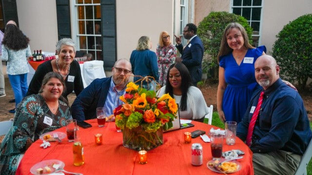 A group of six adults, smiling and seated around a round table with an orange tablecloth, floral centerpiece, and food and drinks, enjoy the Annual Banquet hosted by Phoebe Physicians Group at an outdoor gathering with others in the background.