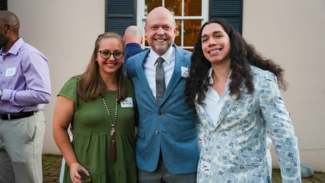 Three people stand outdoors at the Phoebe Physicians Group Annual Banquet, smiling at the camera. The woman on the left wears a green dress and glasses, the man in the middle wears a blue suit, and the man on the right wears a floral blazer.