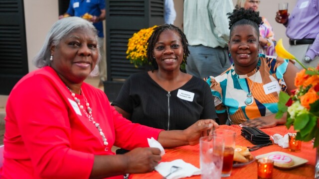 Three women sit smiling at a table decorated with flowers and drinks at the Phoebe Physicians Group Annual Banquet. They wear name tags and colorful outfits, enjoying a lively outdoor social event.