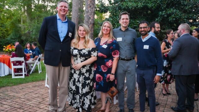 A group of five adults from the Phoebe Physicians Group stands together smiling at an outdoor Annual Banquet, dressed in semi-formal attire. Other guests mingle near tables with orange and white tablecloths, surrounded by trees and greenery.