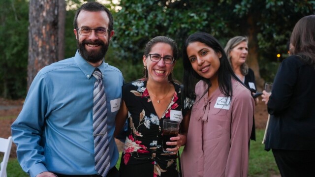 Three people from the Phoebe Physicians Group stand close together and smile at the camera during the Annual Banquet. Dressed in business-casual attire with name tags, they enjoy the outdoor event amid trees and other attendees.