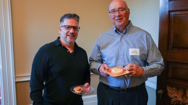 Two men stand indoors, smiling at the camera during the Phoebe Physicians Group Annual Banquet. One holds a plate with a cupcake, while the other shows off a decorated cookie with the number 125 on it. Both wear glasses and casual clothing.