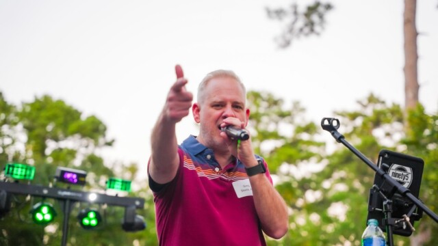 A man in a red striped polo shirt holds a microphone and points toward the camera while performing outdoors at the Phoebe Physicians Group Annual Banquet. Trees and stage equipment are visible in the background.