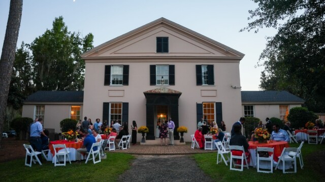 People sit at round tables covered with red and white tablecloths outside a large, light-colored two-story house during the Phoebe Physicians Group Annual Banquet at dusk. Each table features floral centerpieces, creating an inviting outdoor event atmosphere.