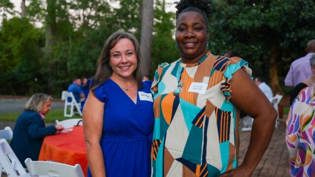 Two women from Phoebe Physicians Group smile and pose together at the Annual Banquet, wearing name tags. One is in a blue dress, the other in a geometric-patterned top. People are seated at tables with trees surrounding them outdoors.