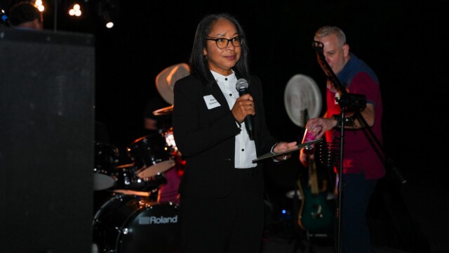 At the Phoebe Physicians Group Annual Banquet, a woman in a black suit and glasses speaks into a microphone outdoors, holding a notepad. Musical instruments and another person with a guitar are visible in the background.