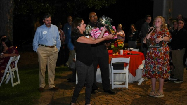 At the Annual Banquet, a woman holding a pink gift basket stands in front of a group at an outdoor nighttime event for Phoebe Physicians Group, as others watch and a woman with a microphone stands nearby. Tables with red tablecloths are seen in the background.