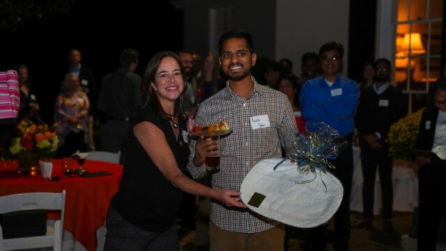 A woman and a man stand smiling at the Phoebe Physicians Group Annual Banquet at night. The man holds a wrapped gift and a drink, while guests in business attire mingle in the background. Both wear name tags.