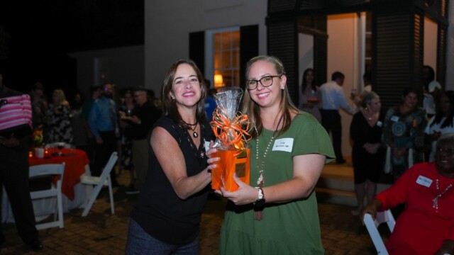 Two women from the Phoebe Physicians Group smile and hold a cellophane-wrapped gift with an orange bow at the Annual Banquet, surrounded by people in front of a building at night.