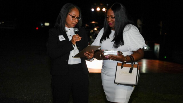Two women stand together at the Annual Banquet hosted by Phoebe Physicians Group; one in a black suit holds a microphone and clipboard, while the other in a white dress holds a purse and plate, both engaged in conversation.