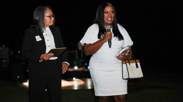 Two women stand together at the Phoebe Physicians Group Annual Banquet, held outdoors at night. One, in a white dress, speaks into a microphone and holds a purse; the other, in a black suit, holds a notepad and looks at her.