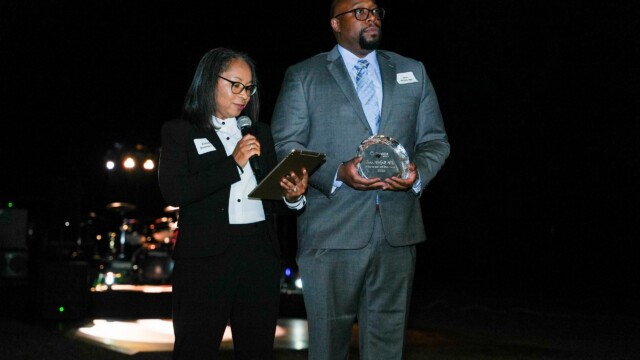 At the Phoebe Physicians Group Annual Banquet, a woman in a black suit speaks into a microphone while holding a clipboard, standing next to a man in a gray suit and blue tie holding a glass award outdoors at night before a dimly lit stage.