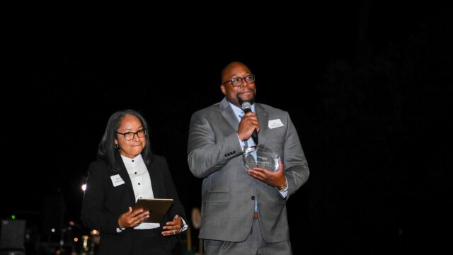 At the Phoebe Physicians Group Annual Banquet, a man in a gray suit speaks into a microphone while holding a glass award. A woman in a black suit and glasses stands beside him with a clipboard. Both face an audience against a dark background.
