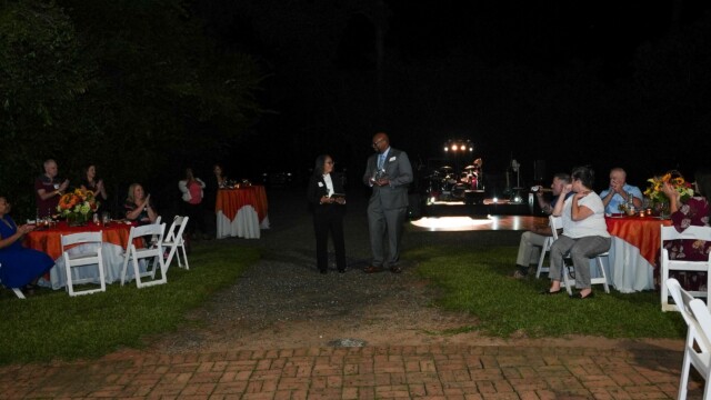 At the Physicians Group Annual Banquet, people sit at round tables with orange tablecloths outdoors at night, watching two walk together near a lit dance floor and a stage with performers in the background.