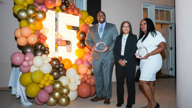 Three people pose indoors beside a large, illuminated 15 sign decorated with balloons at the Phoebe Physicians Group Annual Banquet. Two hold glass awards and smile in business attire, with a door and windows in the background.