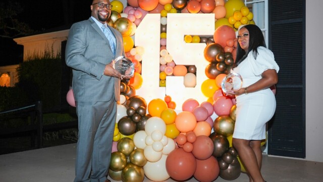 A man in a gray suit and a woman in a white dress smile while holding awards at the Phoebe Physicians Group Annual Banquet, standing in front of a large illuminated 15 surrounded by colorful balloons.