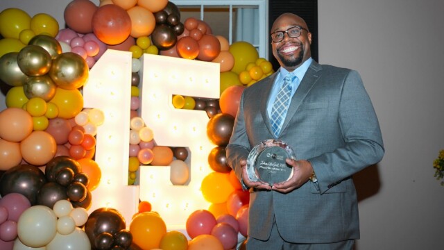 A man in a gray suit and glasses smiles while holding an award at the Phoebe Physicians Group Annual Banquet. Behind him is a large, illuminated 15 surrounded by colorful balloons in yellow, orange, gold, and pink tones.