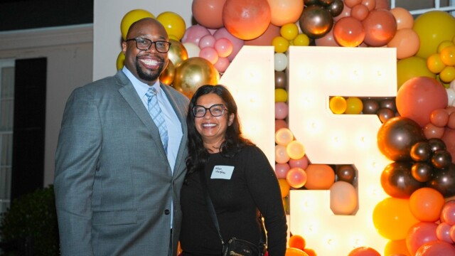 A man in a gray suit and a woman wearing glasses and a black top smile in front of a large, illuminated 15 sign with colorful balloons at the Physicians Group Annual Banquet.