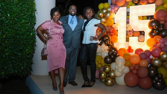 Three people pose and smile in front of a large illuminated “15” sign surrounded by colorful balloons at night, celebrating the Annual Banquet for Phoebe Physicians Group. Two women stand on either side of a man, all dressed in semi-formal attire.