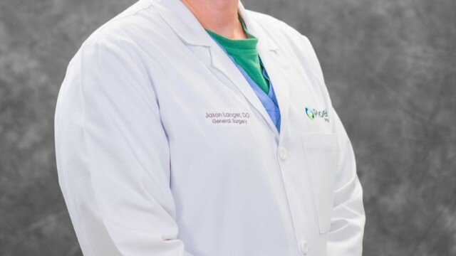 A man wearing a white lab coat stands in front of a gray textured background, smiling at the camera with his hands clasped in front of him—Phoebe Surgeon, known for using his gaming hobby to help children. A man in a white lab coat stands before a gray studio backdrop, smiling with hands clasped, inspired by Phoebe's mission to help kids.
