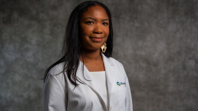 A woman with long dark hair wearing a white lab coat and a white top stands in front of a gray backdrop, smiling gently at the camera, celebrating National Primary Care Week and inspiring new physicians. A woman with long dark hair wearing a white lab coat and a white top stands in front of a gray backdrop, smiling gently at the camera, celebrating National Primary Care Week and inspiring new physicians.