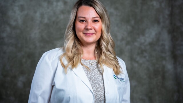 A woman with long blonde hair wearing a white lab coat and patterned shirt stands in front of a gray mottled background, smiling slightly at the camera, representing new physicians during National Primary Care Week. A woman with long blonde hair wearing a white lab coat and patterned shirt stands in front of a gray mottled background, smiling slightly at the camera, representing new physicians during National Primary Care Week.