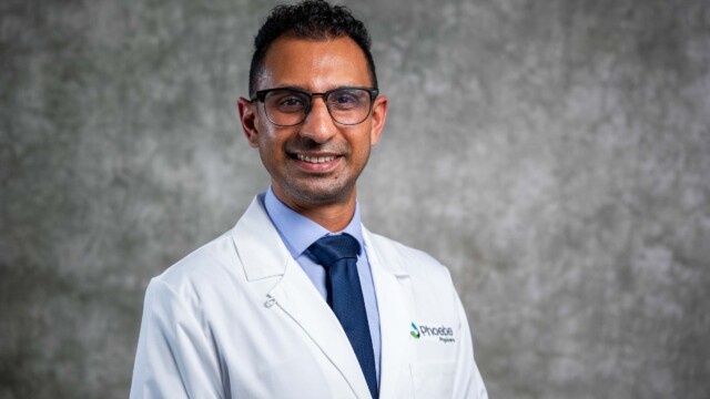 A man with short dark hair and glasses, wearing a white lab coat and blue tie, stands in front of a textured gray background, smiling at the camera—an inspiring image for National Primary Care Week and new physicians. A man with short dark hair and glasses, wearing a white lab coat and blue tie, stands in front of a textured gray background, smiling at the camera—an inspiring image for National Primary Care Week and new physicians.