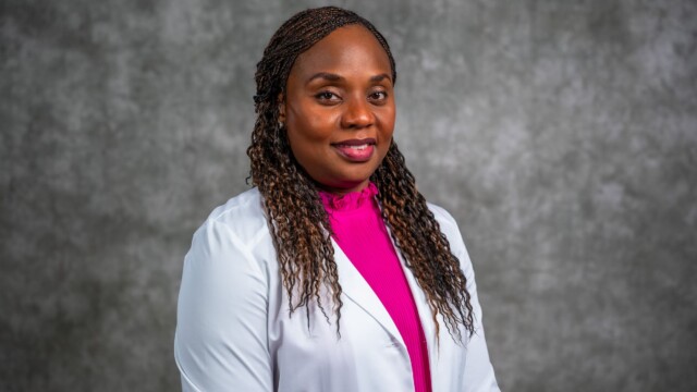 A woman with long braided hair wearing a white lab coat over a bright pink top stands in front of a gray textured background, smiling slightly at the camera—an inspiring image for National Primary Care Week and new physicians. A woman with long braided hair wearing a white lab coat over a bright pink top stands in front of a gray textured background, smiling slightly at the camera—an inspiring image for National Primary Care Week and new physicians.
